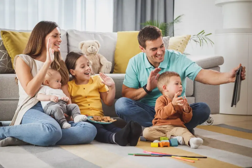 A family on their living-room rug waving at a tablet during a video call with relatives